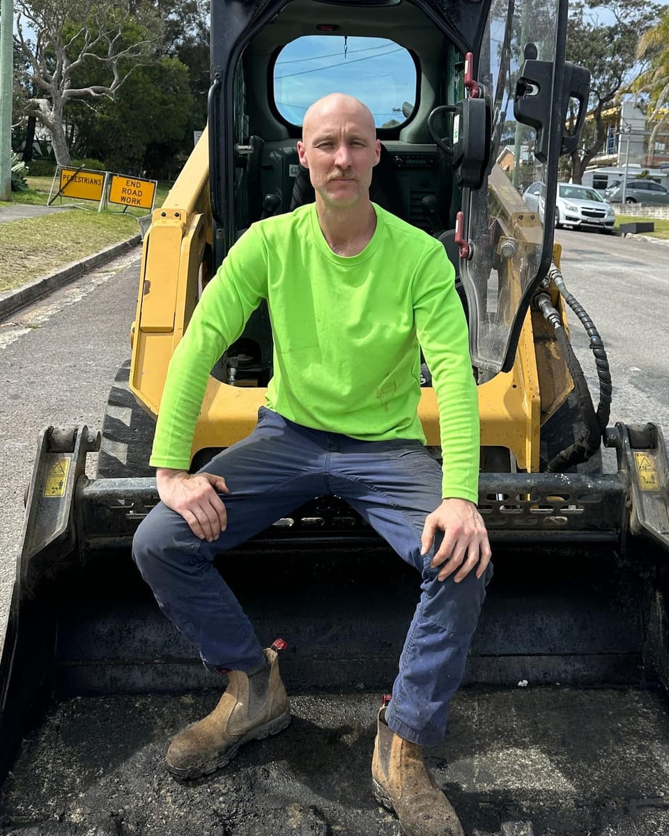 Nathan, founder of All Trade Web & SEO, sitting in the bucket of a skid steer loader in a hi-vis work shirt